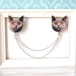 May include: A decorative brooch featuring two cat faces with orange eyes, connected by a silver chain. The cat faces are gray and white, and the brooch has a silver-colored metal finish. The brooch is displayed on a white frame.