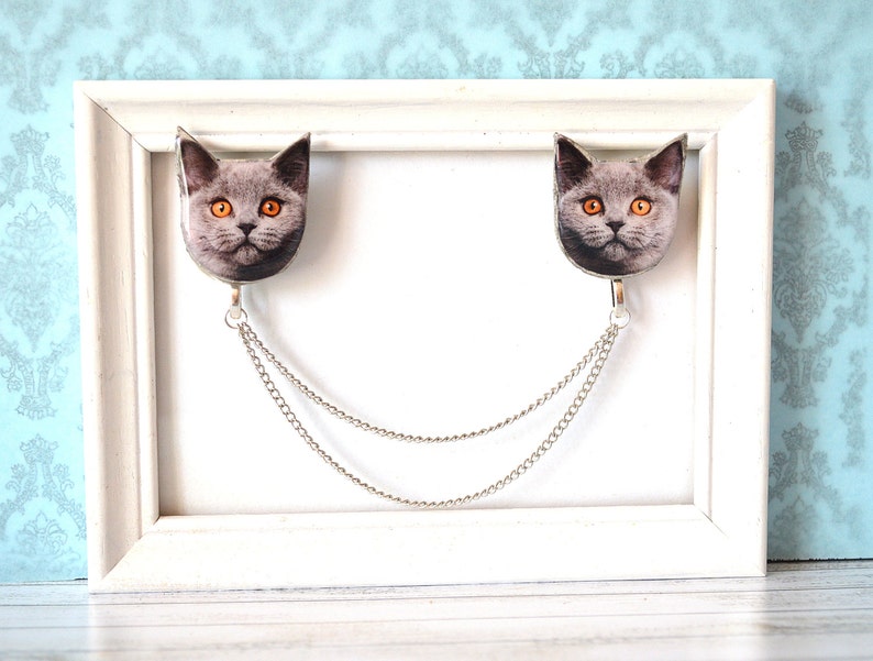 May include: A decorative brooch featuring two cat faces with orange eyes, connected by a silver chain. The cat faces are gray and white, and the brooch has a silver-colored metal finish. The brooch is displayed on a white frame.