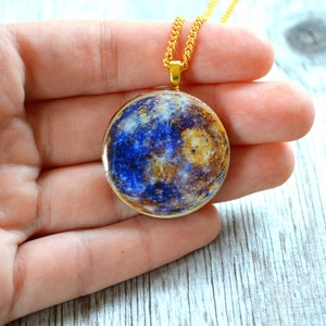 May include: A gold-toned necklace featuring a round pendant with a planetary design in shades of blue, brown, and gold. The pendant is suspended from a delicate gold chain. The necklace is held in a hand.