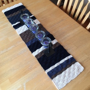 May include: A knitted table runner with stripes of white, brown, and blue. The runner is decorated with a metal candle holder and five purple candles.