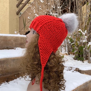 May include: Red knitted hat with a long, dangling tail and a gray faux fur pom pom. The hat is on a mannequin head with long brown hair.