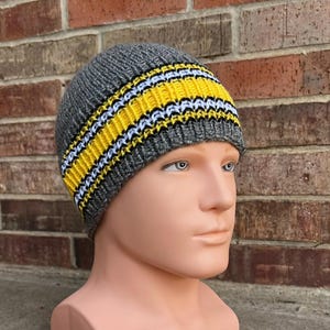 Op de afbeelding: Een grijze gebreide muts met een geel en wit gestreepte band. De hoed wordt getoond op een mannequinhoofd. Een winteraccessoire.
