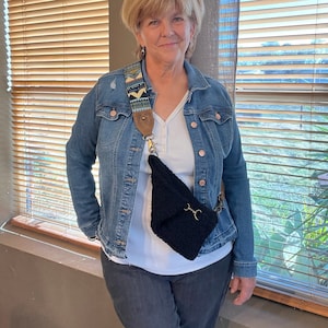 May include: A woman wearing a denim jacket, a white shirt, and dark blue jeans. She is carrying a black crocheted crossbody bag with a brown and blue patterned strap.