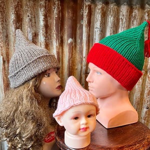 May include: Three knitted elf hats in different colors: brown, pink, and green with red trim. The hats have pointed tops and are displayed on mannequin heads.