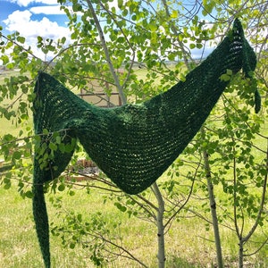 Könnte beinhalten: Ein grüner gestrickter Schal, der an einem Baumzweig hängt. Der Schal ist aus weichem, strukturiertem Garn gefertigt und hat eine dreieckige Form.