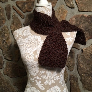 Puede incluir: Una bufanda de punto marrón expuesta en un maniquí blanco con un patrón de paisley. La bufanda está hecha de un hilo suave y texturizado y tiene un diseño simple y elegante.