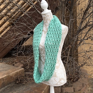 May include: A light green crocheted infinity scarf on a white mannequin. The scarf is made with a simple stitch pattern and has a loose, drapey feel.