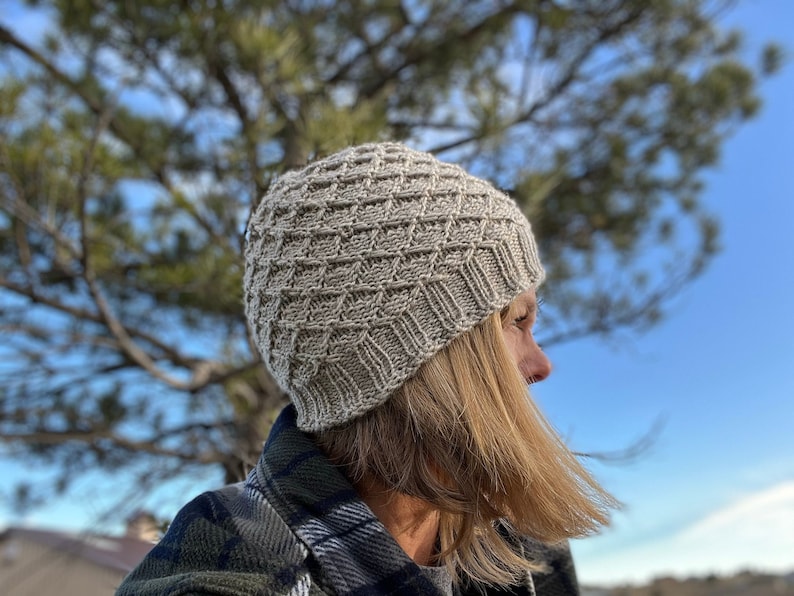 May include: A beige knitted hat with a textured pattern. The hat is worn by a person with long, blonde hair. The person is standing outdoors with a tree in the background.
