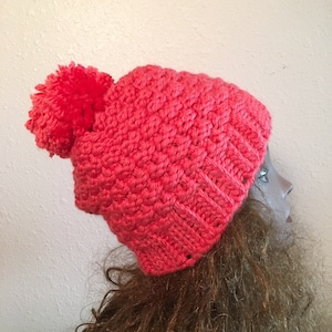 Puede incluir: Un gorro de punto de color rosa brillante con un pompón esponjoso en la parte superior.