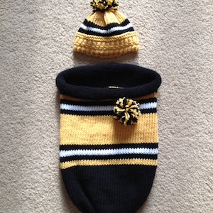 Puede incluir: Un saco de dormir para bebé de punto a rayas negro y dorado con un gorro a juego de punto a rayas negro y dorado con un pompón blanco.