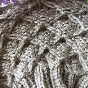 May include: Close-up of a beige knitted fabric with a textured pattern. The fabric is being worked on a loom.