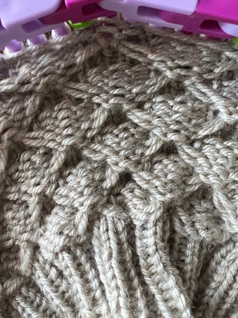 May include: Close-up of a beige knitted fabric with a textured pattern. The fabric is being worked on a loom.