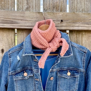 May include: A pink knitted scarf with a knotted design, hanging on a wooden fence.