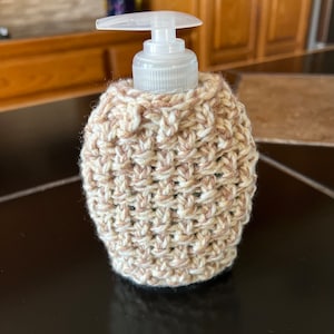 May include: A crocheted soap dispenser cover in beige and white. The cover is made with a textured stitch pattern.