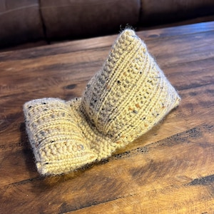 May include: A hand-knitted book or tablet stand in beige. The stand is made from textured yarn with coloured flecks. It is folded into a triangular shape to support a book or tablet. The stand is placed on a wooden surface.