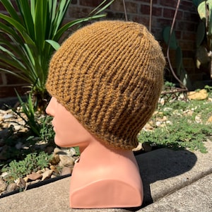 May include: A knitted beanie in a warm, golden-brown color. The hat is made of thick yarn, with a ribbed texture. The beanie is displayed on a mannequin head, showcasing its shape and fit. This winter hat is a cozy accessory.