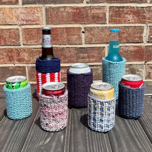 May include: A collection of handcrafted beverage insulators in various colors and patterns. The image features several knitted cozies in shades of blue, purple, and a red, white, and blue design. Some are holding cans and bottles.