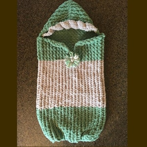 May include: A green and white crocheted baby sleeping bag with a hood. The sleeping bag has a flower embellishment.