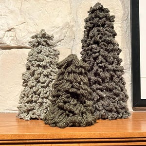Puede incluir: Tres árboles de Navidad hechos a mano con ganchillo en varios tamaños y tonos de gris y verde. Los árboles se exhiben sobre una superficie de madera frente a una pared de piedra. Los árboles tienen un aspecto texturizado y rústico.