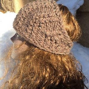 Puede incluir: Un gorro de punto marrón con un agujero para la coleta. El gorro está hecho de un hilo grueso y texturizado.