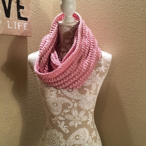 May include: A light pink, chunky knit infinity scarf displayed on a mannequin. The scarf is made of a soft, textured yarn and is draped around the mannequin's neck. The mannequin is patterned with a floral design. A sign in the background reads "Happiness, Love, Life."