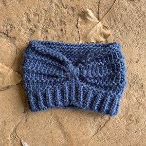 Op de afbeelding: Een blauwe gebreide hoofdband met een geknoopt detail. De hoofdband is gemaakt van zacht, getextureerd garen.