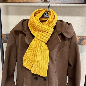 May include: A mustard yellow knitted scarf draped over a brown coat. The scarf has a ribbed texture and is made of a soft yarn. The coat has a classic design with a notched collar and two buttons. The scarf is hanging on a black hanger.
