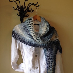 May include: A blue and white crocheted shawl with a lacy pattern. The shawl is draped over a white denim jacket and hanging on a black metal hook.