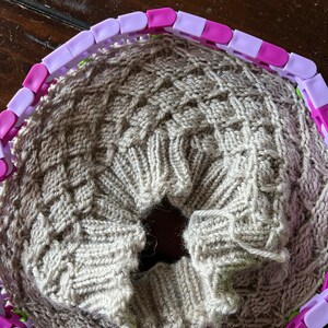 May include: A beige knitted fabric is being worked on using a circular knitting loom with purple and white plastic pieces.