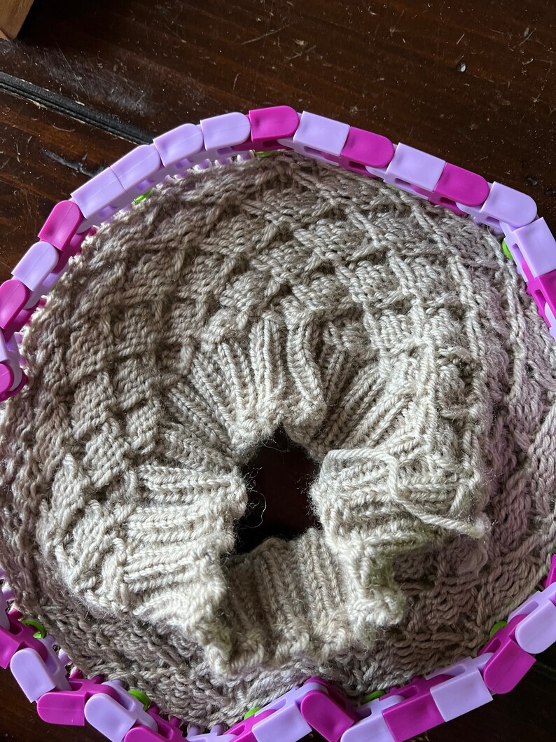 May include: A beige knitted fabric is being worked on using a circular knitting loom with purple and white plastic pieces.