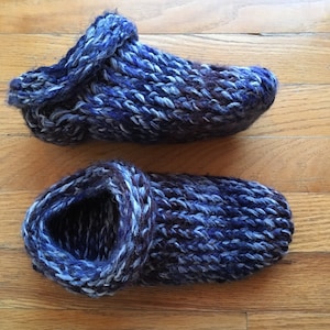 May include: Two handmade knitted slippers in shades of blue, gray, and purple sit on a wooden floor. The slippers have a thick, textured appearance, with a folded-over cuff and a closed toe design. The yarn appears soft and warm.