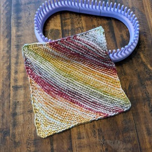 May include: A square, hand-knitted item with a diagonal striped pattern in shades of red, yellow, green, and white. A purple knitting loom is in the background. The knitted item appears to be a washcloth or small blanket.