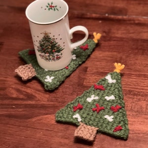 Puede incluir: Dos posavasos de árbol de Navidad de crochet verde con detalles rojos y blancos. Un posavasos tiene una taza blanca con un diseño de árbol de Navidad.