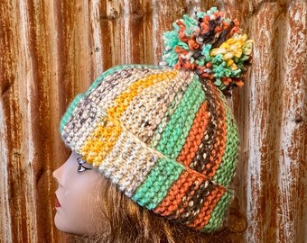 May include: A striped knit beanie with a multi-colored pom pom. The beanie is made with a variety of colors, including beige, orange, green, and brown.