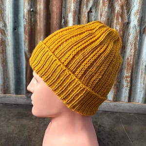 May include: Mustard yellow knit beanie hat. The hat is ribbed and has a folded brim. The hat is displayed on a mannequin head. The background is a corrugated metal wall.