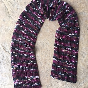 Puede incluir: Bufanda tejida a mano con un patrón de canalé. La bufanda presenta una mezcla de colores, incluyendo negro, burdeos, blanco y motas doradas y rosas. Hecha de hilo suave, está diseñada para brindar calidez y estilo.