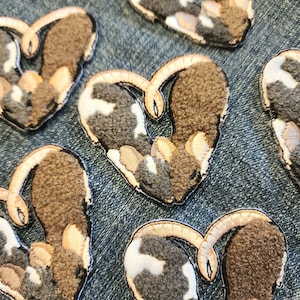 May include: Heart-shaped embroidered patches with a textured, plush appearance. The patches feature a mix of brown, gray, and white colors, with a cream-colored border. These patches are likely for clothing or craft projects.