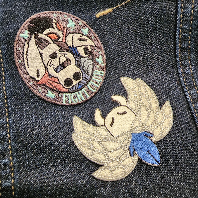 May include: Two embroidered patches on a blue denim jacket. The first patch is round and features a white, blue, and pink creature with the text "Fight Club". The second patch is a white, blue, and silver creature with wings.