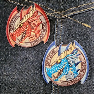 May include: Two embroidered patches featuring dragons with the text "Official Dragon Rider" on a denim background. The first patch features a red dragon with brown accents. The second patch features a blue dragon with brown accents.
