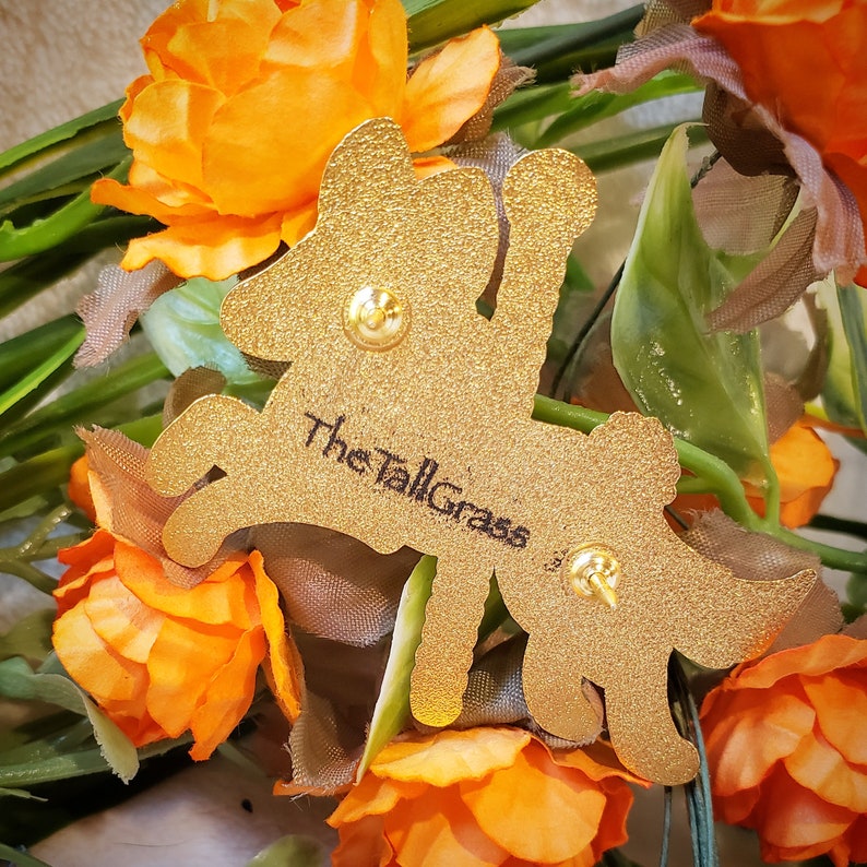K&ouml;nnte beinhalten: Goldfarbener Metallstift mit einem stilisierten Hirsch und dem Text "The Tall Grass" auf der Seite. Der Stift ist von orangefarbenen Stoffblumen umgeben.