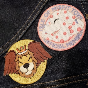 May include: Two embroidered patches. One is pink with white polka dots and says "Isle of Misfit Toys Official Member". The other is gold with a brown and white cartoon dog wearing a crown and says "Isle of the Misfits".