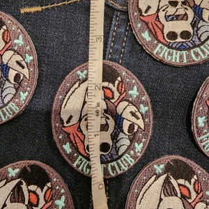 May include: Close-up of five embroidered patches featuring a character from the video game Hollow Knight. The patches are circular and have a dark purple background with a light blue border. The character is depicted in a white and gray color scheme with red accents. The text "FIGHT CLUB" is embroidered in a light blue font on the bottom of each patch. A ruler is visible in the background, measuring 2 inches.