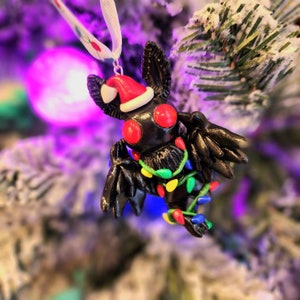 Könnte beinhalten: Ein schwarzer, fledermausförmiger Weihnachtsschmuck mit roten Augen und einem Weihnachtsmannhut. Der Schmuck ist mit einer Lichterkette in bunten Farben geschmückt. Er hängt an einem weißen Band an einem verschneiten Weihnachtsbaum.