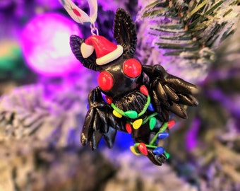 May include: A black bat-like Christmas ornament with red eyes and a Santa hat. The ornament is decorated with a string of colorful Christmas lights. It is hanging from a white ribbon on a snowy Christmas tree.