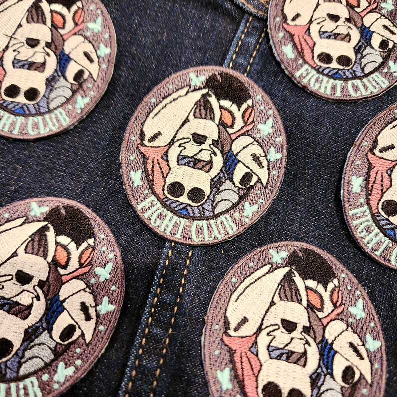 May include: Five embroidered patches featuring a white character with a black mask and a pink cape. The character is holding a sword and a shield. The patches are circular and have a brown background with the text "Fight Club" in white. The patches are arranged in a circle on a blue denim background.