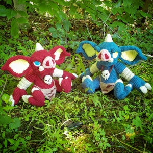 May include: Two crocheted creature dolls, one deep red and one blue, each featuring large pointed ears with pale yellow centers, a snout with a pink nose, a single horn on the head, and a skull pendant hanging from a dark cord around the neck.