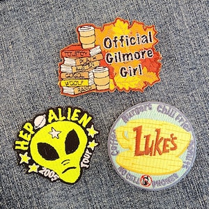May include: Three embroidered patches, one yellow with a green alien head and the text "HEP ALIEN TOUR 2005", one yellow with a brown coffee cup and the text "Luke's", and one orange with the text "Official Gilmore Girl".
