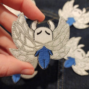 May include: A white and silver embroidered patch of a character with blue wings and a blue body. The character has a white face with black eyes and a black mouth.