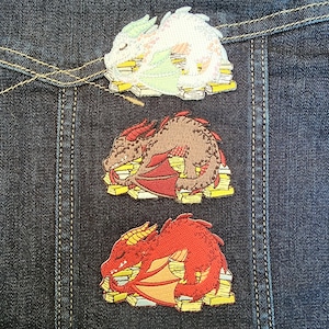 May include: Three embroidered dragon patches in white, brown, and red, each resting on a pile of yellow and white books. The patches are on a dark blue denim background with visible stitching.