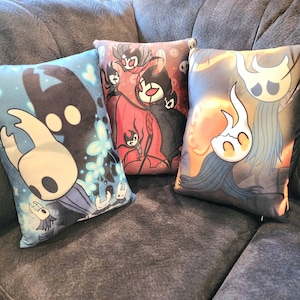 Può includere: Tre cuscini decorativi con sfondo grigio scuro e disegni grafici colorati con personaggi del videogioco Hollow Knight. I cuscini sono realizzati in tessuto morbido e hanno una sensazione confortevole.